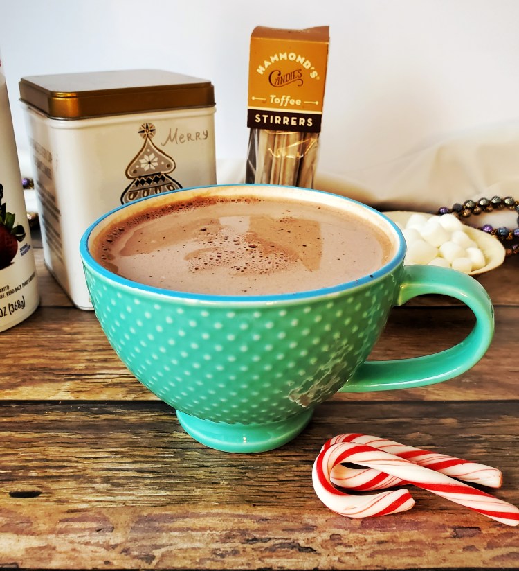 Hot cocoa in a teal cup. a small bowl of mini marshmallows is on the right, a tin of cocoa mix, Hammond's Toffee Stir sticks, and a can of whipped cream. A couple of candy canes are in the foreground.
